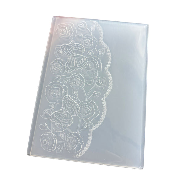 A rectangular translucent plastic mold or stamp with an etched floral rose pattern and scalloped border, shown on a plain white background.