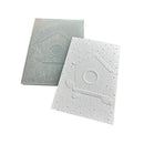 A white embossed card or plaque featuring a birdhouse design with pine branches and a circular window, shown in front of a translucent matching backing sheet on a plain white background.