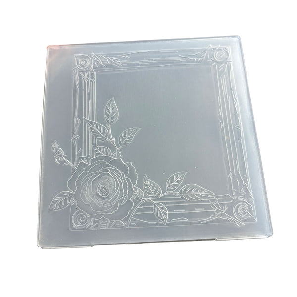 A square translucent plastic mold or stamp with an embossed floral design featuring a rose and leaves in the bottom-left corner, set against a plain white background.