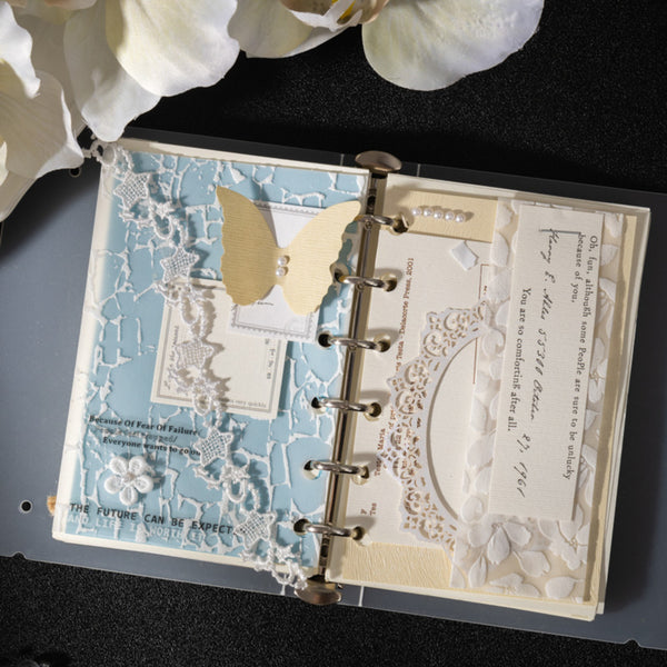 Open scrapbook with decorative pages and a butterfly on a dark surface with white flowers.