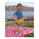 Young girl holding a yellow bag with an orange design on a red and white checkered blanket by a lake.