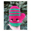 Hand holding Crocheted pink and green mitten amigurumi with a embodied smiley face front of a Christmas tree.