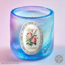 Candle in a blue glass holder with a floral label on a light purple background