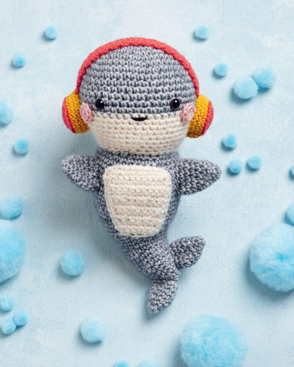 Crocheted shark toy with headphones on a light blue background with colorful pom-poms.