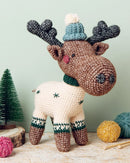 Crocheted moose toy with a hat and scarf on a wooden surface with yarn balls and a small tree.