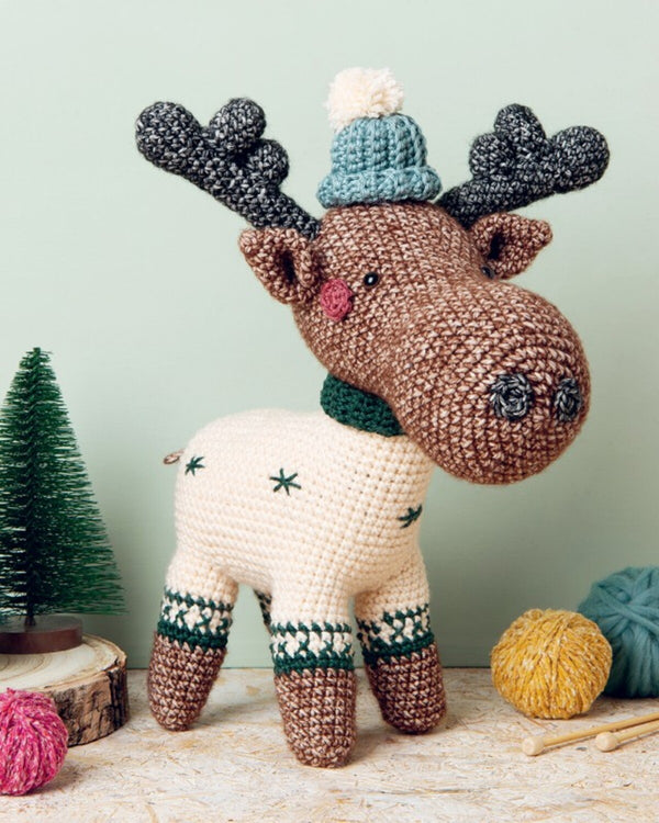 Crocheted moose toy with a hat and scarf on a wooden surface with yarn balls and a small tree.