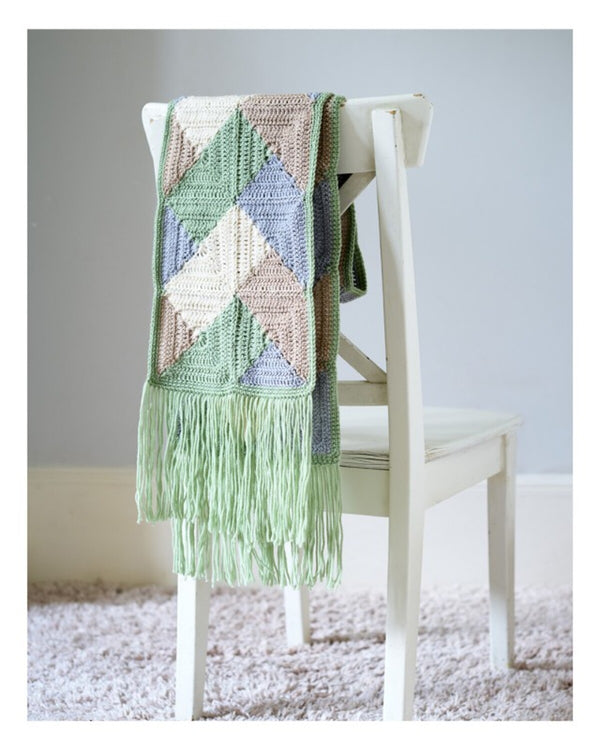 Crocheted blanket with geometric pattern and fringe on a white chair against a light gray wall.