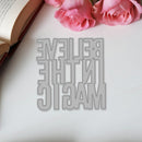 Metallic bookmark with 'Believe in the Magic' text on a white surface with pink roses and an open book.