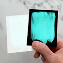Turquoise color swatch on a black card held by a hand against a marble background