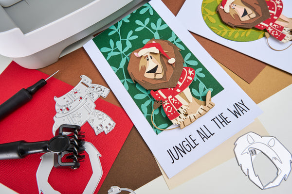 Card with a lion wearing a Santa hat and 'Jungle All The Way' text, surrounded by crafting tools on a wooden surface.