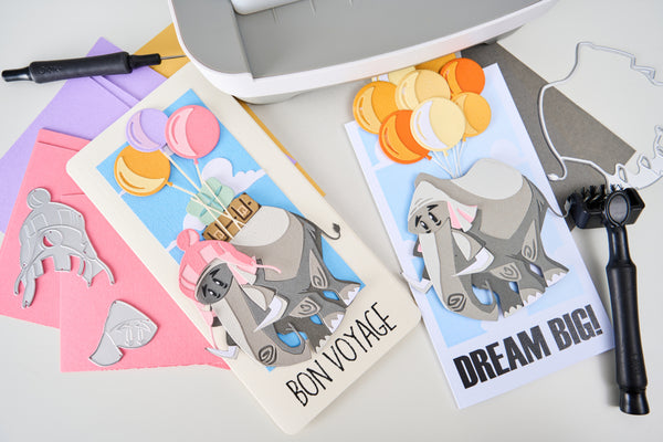 Two greeting cards with elephant designs and text, surrounded by crafting tools on a light surface.