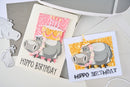 Two hippo-themed birthday cards on a gray surface with a laptop in the background.