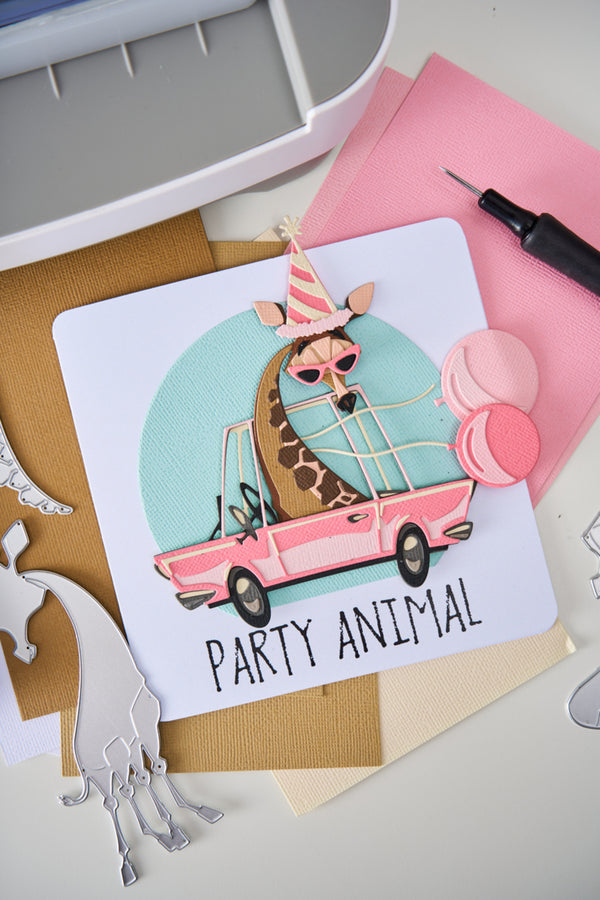 Card with a giraffe in a car wearing sunglasses and a party hat, surrounded by crafting supplies.