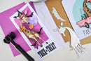 Halloween-themed greeting card with a giraffe in a witch costume on a purple background, surrounded by other cards and a die-cut animal.