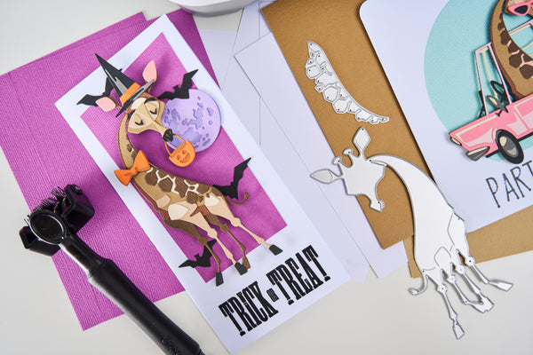 Halloween-themed greeting card with a giraffe in a witch costume on a purple background, surrounded by other cards and a die-cut animal.