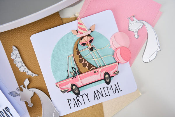 Card with a giraffe in a party hat and sunglasses driving a pink car, surrounded by die-cut shapes on a light background.