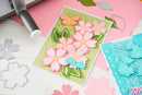 Card with pink flowers and butterfly on a pink surface with crafting tools.