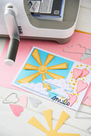 Card with sun and cloud design on pink surface with crafting tools