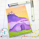Card with Easter design featuring crosses and 'he is risen' text, placed on a grid surface with stamping tools.
