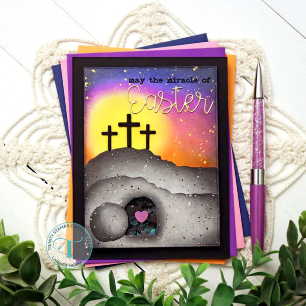 Easter card with crosses and a heart on a colorful background, placed on a textured surface with a pen and plants.
