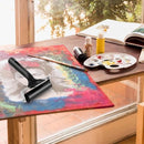 Artistic setup with a roller, paint bottles, and a palette on a table by a window.