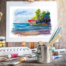Artistic setup with a framed watercolor painting of a coastal scene, paint supplies, and a metal container on a wooden surface.