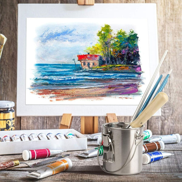Artistic setup with a framed watercolor painting of a coastal scene, paint supplies, and a metal container on a wooden surface.