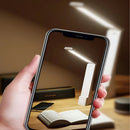 Hand holding a smartphone with a screen displaying a desk setup with a lamp and book.