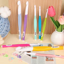 Set of colorful crochet hooks on a wooden surface with a vase of flowers and yarn.