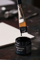 Jar of Absolute Black paint with a brush dipping into it on a dark surface.