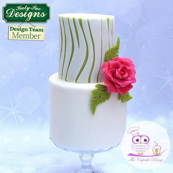 Two-tiered cake with green and white design and pink rose on a light blue background, featuring Katy Sue Designs branding.