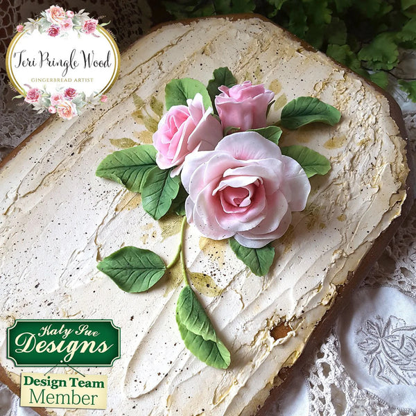 Decorative cake with pink roses and green leaves on a textured surface, featuring Katy Sue Designs branding.